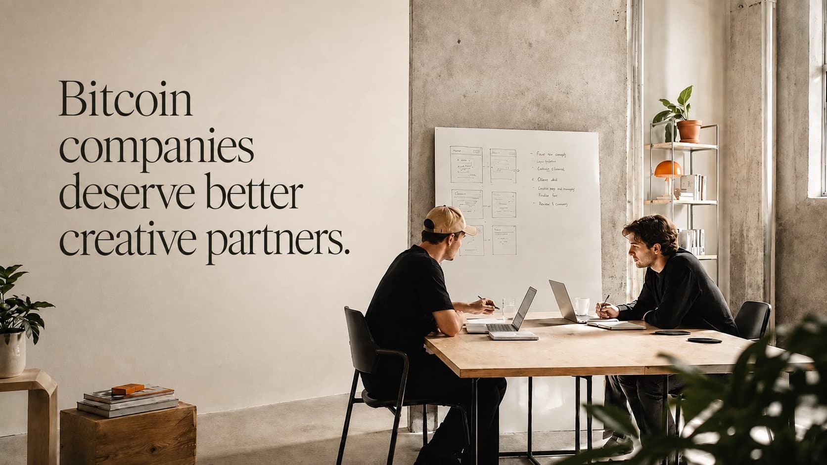 A studio wall reading Bitcoin companies deserve better creative partners, with two operators working at a table in front of a whiteboard of wireframes